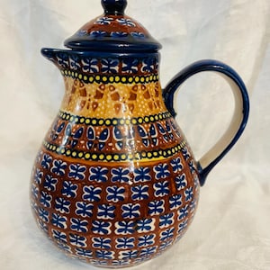 May include: A ceramic pitcher with a lid, featuring a rounded body and a curved handle. The surface is decorated with a repeating pattern of small blue floral shapes on a brown background, accented by yellow and blue bands around the neck.