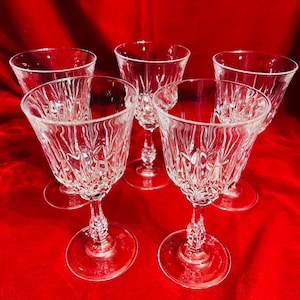 May include: Five clear crystal wine glasses with intricate etched designs. Each glass features a round base, a detailed stem, and a flared bowl. The glasses are arranged on a red background, showcasing their elegant details.