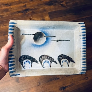 May include: Rectangular ceramic serving dish with a hand-painted design. The dish features a blue and white color scheme with a stylized moon and three dark figures. The rim is decorated with blue vertical lines.
