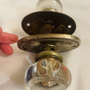 May include: A pair of vintage glass doorknobs with a clear, faceted design. The knobs are attached to aged metal plates and hardware, showing signs of wear. The hardware is likely made of brass or a similar metal. The overall aesthetic is antique or retro.