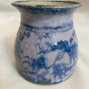 May include: A handmade ceramic vase with a rounded body and a slightly flared rim. The vase features a mottled blue and lavender glaze with darker blue accents. The interior is a light beige color. The vase is approximately 5 inches tall.