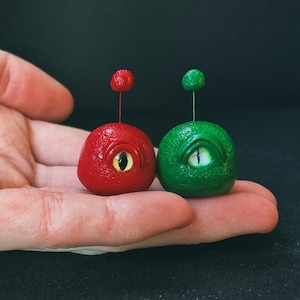 May include: Two small, handmade, ceramic ornaments with a single eye. One is red with a black eye and the other is green with a white eye.