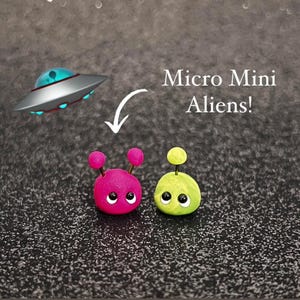 Pode incluir: Duas figuras alienígenas em miniatura, uma rosa com antenas e outra verde limão com uma única antena, são apresentadas. O texto "Micro Mini Aliens!" é exibido acima das figuras, com um OVNI de desenho animado no fundo.