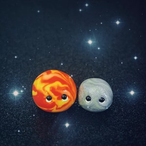 Puede incluir: Dos pequeños planetas con ojos saltones, uno es naranja y amarillo con remolinos y el otro es gris con un remolino blanco. Los planetas están sobre un fondo azul oscuro con estrellas blancas.