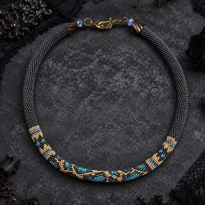 May include: A circular beaded necklace featuring a predominantly black base with a repeating geometric pattern in metallic gold and turquoise blue. The necklace is finished with gold-toned metal end caps and a decorative gold-toned hook clasp.