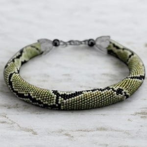 May include: A beaded bracelet with a snake-like pattern. The bracelet features a green and black design, with a clasp closure. The beads are small and tightly woven, creating a textured surface. The bracelet is a unique accessory.