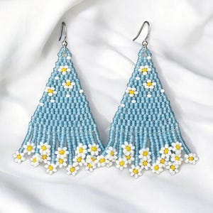 May include: A pair of blue beaded earrings featuring a daisy pattern. The earrings are triangular, with a beaded fringe at the base. The daisies have white petals and yellow centres. They hang from silver-coloured hooks.