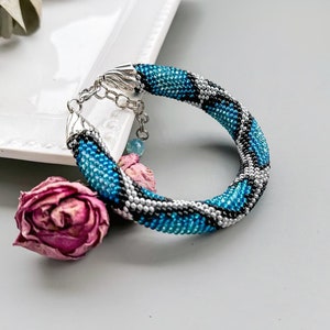 May include: A blue and silver beaded bracelet with a snake skin pattern. The bracelet has a silver clasp and is lying on a white surface.
