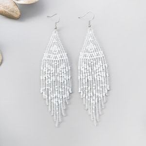 Puede incluir: Un par de pendientes blancos de cuentas con forma de lágrima. Los pendientes tienen un gancho plateado y están hechos de pequeñas cuentas blancas. Se exhiben sobre una superficie gris claro con flores y hojas secas.