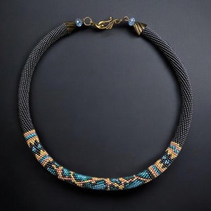Pode incluir: Um colar de contas pretas com um padrão geométrico em tons de azul, dourado e laranja. O colar tem um fecho de bronze com dois pequenos cristais azuis.
