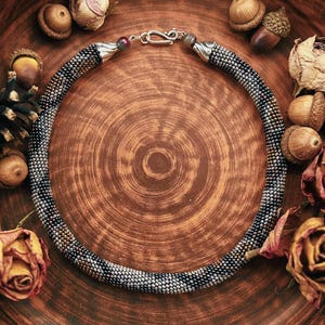 May include: A beaded necklace with a silver clasp rests in a brown ceramic bowl, surrounded by dried roses, acorns, and a pinecone. The bowl sits on a surface with vintage paper and the word "Autumn" visible.