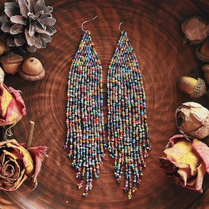 Puede incluir: Un par de pendientes largos de cuentas multicolores, presentados en un cuenco de madera rústico con rosas secas, bellotas y papel vintage. Los pendientes son el punto focal, mostrando un estilo bohemio.