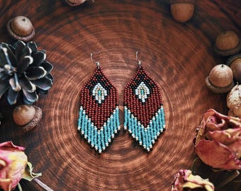 May include: Handmade beaded earrings in a teardrop shape, featuring a pattern of red, turquoise, and white beads. The earrings are displayed in a rustic brown bowl with dried flowers, acorns, and vintage paper accents. The earrings are approximately 3 inches long.