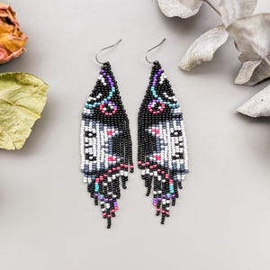 Puede incluir: Un par de pendientes de cuentas con un patrón geométrico negro, blanco, rosa y azul. Los pendientes tienen flecos en la parte inferior y se exhiben sobre una superficie gris con flores secas.
