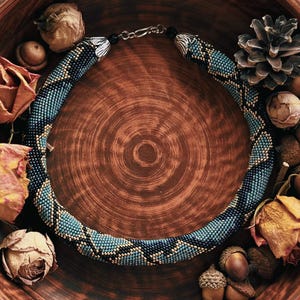 Pode incluir: Um colar de contas com um padrão geométrico azul e preto repousa numa tigela de cerâmica castanha. A tigela é rodeada por rosas secas, bolotas e papel vintage. Um pinha e outros elementos decorativos completam o arranjo.