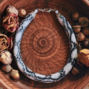 May include: A beaded necklace with a black and white pattern rests in a large, brown, ceramic bowl. The bowl is surrounded by dried roses, acorns, and vintage paper. The necklace is approximately 46 cm in length.