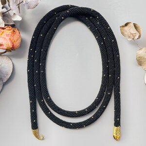 May include: A long, flexible rope necklace crafted from tiny black beads with subtle gold-colored accents throughout. The necklace features two textured, gold-toned metal end caps that are shaped like stylized, curved serpent or animal heads.
