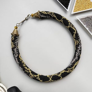 May include: A black, gold and silver beaded necklace with a geometric pattern. The necklace is made of small beads and has a clasp closure.