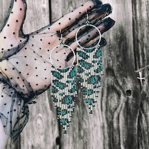 Könnte beinhalten: Ein Paar lange, geperlte Ohrringe mit einem Ringdesign. Die Ohrringe sind aus schwarzen, weißen und türkisfarbenen Perlen gefertigt. Die Ohrringe hängen an einer Hand, die einen schwarzen Spitzenhandschuh trägt.
