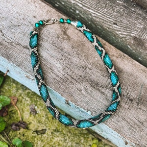Pode incluir: Um colar de contas com um padrão de pele de cobra em tons de verde e preto. O colar tem um fecho prateado.