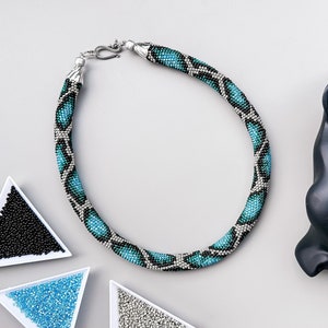 Pode incluir: Um colar de contas turquesa e prata com um padrão de pele de cobra. O colar está sobre uma superfície branca com pequenas tigelas de contas no fundo.