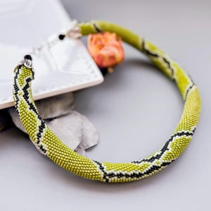 May include: A tubular beaded necklace featuring a vibrant lime green base with intricate black and white geometric patterns. The necklace is finished with silver-toned metal end caps and a clasp mechanism, showcasing a uniform, textured beadwork design.