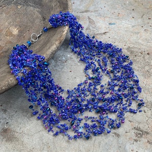 Op de afbeelding: Een meerrijige ketting gemaakt van blauwe en paarse kralen. De ketting is gemaakt van verschillende maten en vormen kralen en is geregen op een zilveren draad.