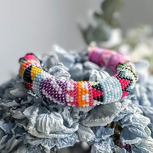 May include: A colorful beaded bracelet with a geometric pattern, resting on a bed of dried hydrangeas.