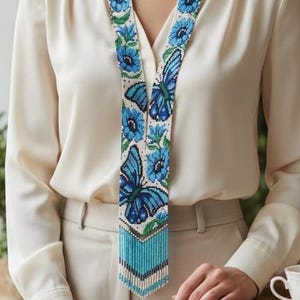 Op de afbeelding: Een kralenketting met een vlinder- en bloemenpatroon in blauw, wit en groen. De ketting wordt gedragen over een crèmekleurige blouse en gecombineerd met een beige broek. De ketting heeft een franje aan de onderkant.