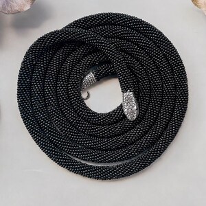 Op de afbeelding: Een lange, opgerolde ketting gemaakt van kleine, uniforme zwarte kralen. De ketting heeft aan één uiteinde een metalen, zilverkleurige slangenkopsluiting met kleine, heldere kristallen en aan het andere uiteinde een bijpassende zilverkleurige punt.