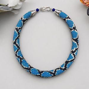 May include: A circular beaded necklace featuring a repeating geometric pattern in bright blue, black, and white seed beads. The necklace is finished with a silver-toned metal hook clasp and small dark blue accent beads at each end of the beaded strand.