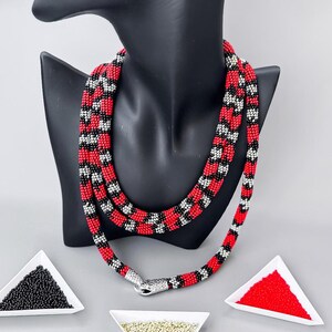 May include: A red, black and silver beaded necklace with a snake clasp on a black mannequin. Three small triangular dishes contain black, silver and red beads.
