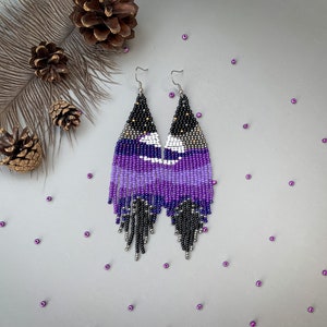 May include: Handmade beaded earrings in black, purple, and white. The earrings have a geometric design with a fringe. They are displayed on a grey surface with pine cones and purple beads. The earrings are approximately 10 cm long.