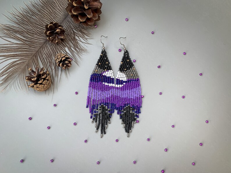 May include: Handmade beaded earrings in black, purple, and white. The earrings have a geometric design with a fringe. They are displayed on a grey surface with pine cones and purple beads. The earrings are approximately 10 cm long.