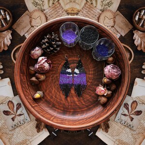 May include: A pair of beaded earrings in a wooden bowl, featuring a black, white, and purple pattern. The earrings are displayed with dried flowers, acorns, and small glasses filled with beads. The image has a rustic, autumnal theme.