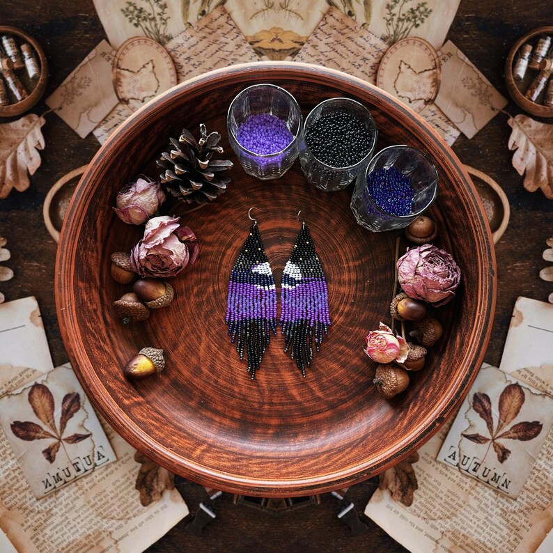 May include: A pair of beaded earrings in a wooden bowl, featuring a black, white, and purple pattern. The earrings are displayed with dried flowers, acorns, and small glasses filled with beads. The image has a rustic, autumnal theme.