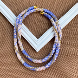 Puede incluir: Un collar de cuentas de doble hebra con un patrón geométrico en tonos de morado, rosa y dorado. El collar está hecho de cuentas pequeñas y tiene un aspecto delicado y femenino.