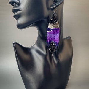 May include: Handmade beaded earrings in a teardrop shape. The earrings feature a gradient of colours, including black, silver, white, and various shades of purple. The earrings are displayed on a black mannequin head.
