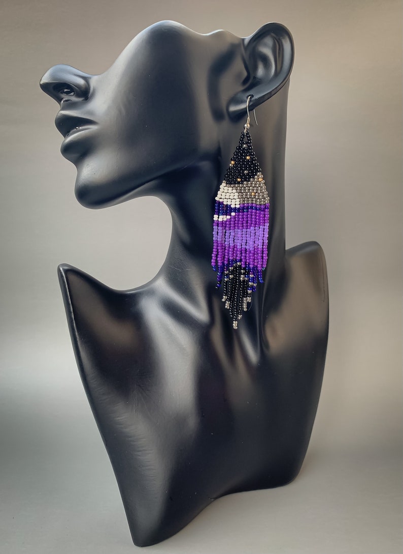 May include: Handmade beaded earrings in a teardrop shape. The earrings feature a gradient of colours, including black, silver, white, and various shades of purple. The earrings are displayed on a black mannequin head.