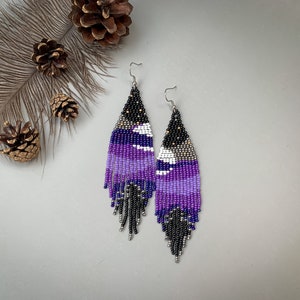 May include: A pair of beaded earrings with a geometric design. The earrings feature a gradient of purple hues, black, white, and silver beads. The earrings have a fringe at the bottom and are suspended from silver hooks.
