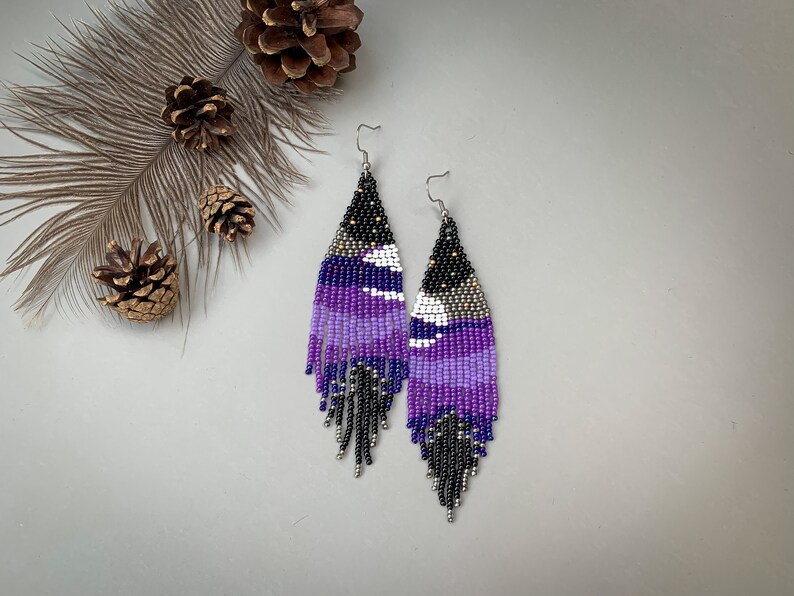 May include: A pair of beaded earrings with a geometric design. The earrings feature a gradient of purple hues, black, white, and silver beads. The earrings have a fringe at the bottom and are suspended from silver hooks.