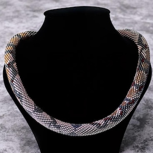 May include: A beaded necklace with a multi-coloured geometric pattern in shades of brown, grey, white, and pink. The necklace is displayed on a black mannequin.
