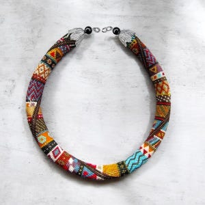 Könnte beinhalten: Eine farbenfrohe Perlenkette mit einem geometrischen Muster in Rot-, Blau-, Gelb- und Brauntönen. Die Halskette hat silberfarbene Verschlüsse und ein detailliertes Design. Ein auffälliges Schmuckstück.