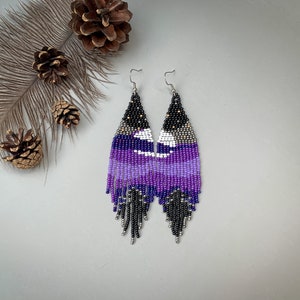May include: Handmade beaded earrings in purple, black, white, and silver. The earrings feature a long, fringed design with a celestial pattern. The earrings are displayed on a light grey surface.