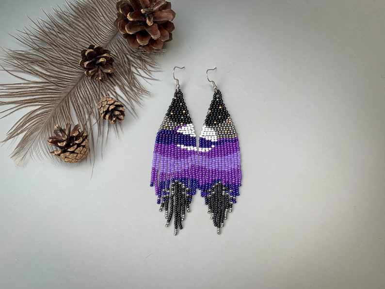 May include: Handmade beaded earrings in purple, black, white, and silver. The earrings feature a long, fringed design with a celestial pattern. The earrings are displayed on a light grey surface.