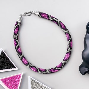 May include: A pink and black beaded necklace with a silver clasp. The necklace is laid out on a white surface with small containers of beads in the background.