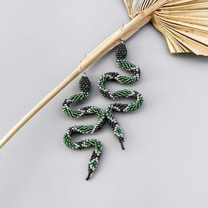 Peut inclure: Une paire de boucles d'oreilles en forme de serpent en perles vertes et noires. Les serpents sont enroulés et ont des accents argentés.