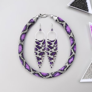 May include: A purple and silver beaded necklace and matching earrings. The necklace is a thick, round cord with a snake skin pattern. The earrings are long and dangle, with a similar pattern to the necklace.