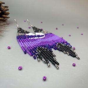 May include: Beaded earrings in a celestial design. The earrings feature a gradient of purple, black, and silver beads, with a white crescent moon shape. The fringe earrings have a silver hook.