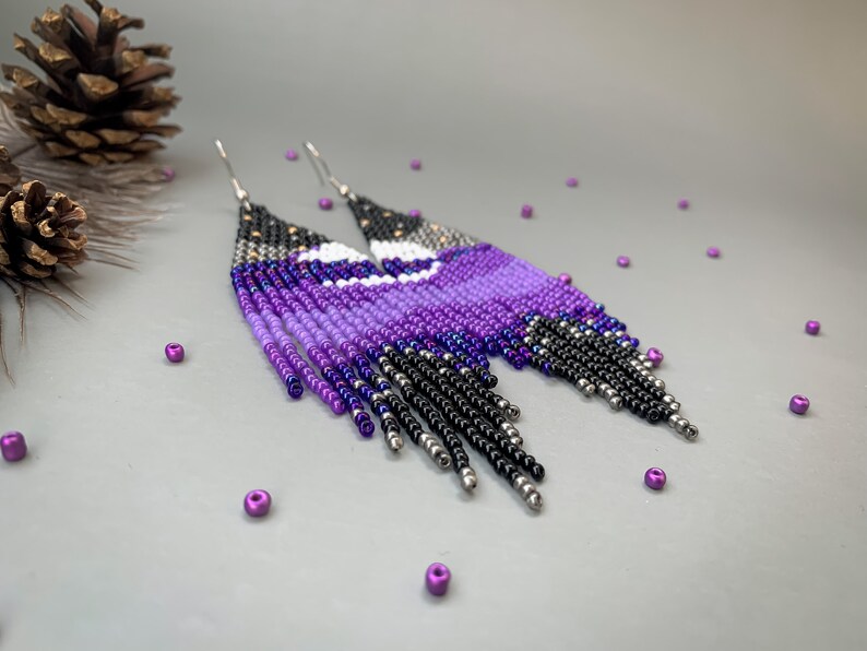 May include: Beaded earrings in a celestial design. The earrings feature a gradient of purple, black, and silver beads, with a white crescent moon shape. The fringe earrings have a silver hook.
